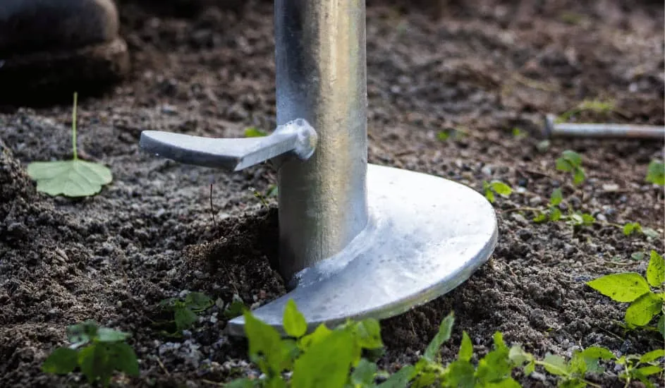 Close-up of a metal screw pile partially embedded in the ground with green plants around.